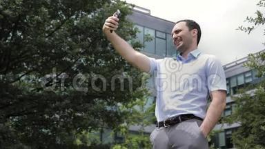 开朗英俊的年轻人微笑着在智能手机相机上自拍。 后台办公大楼